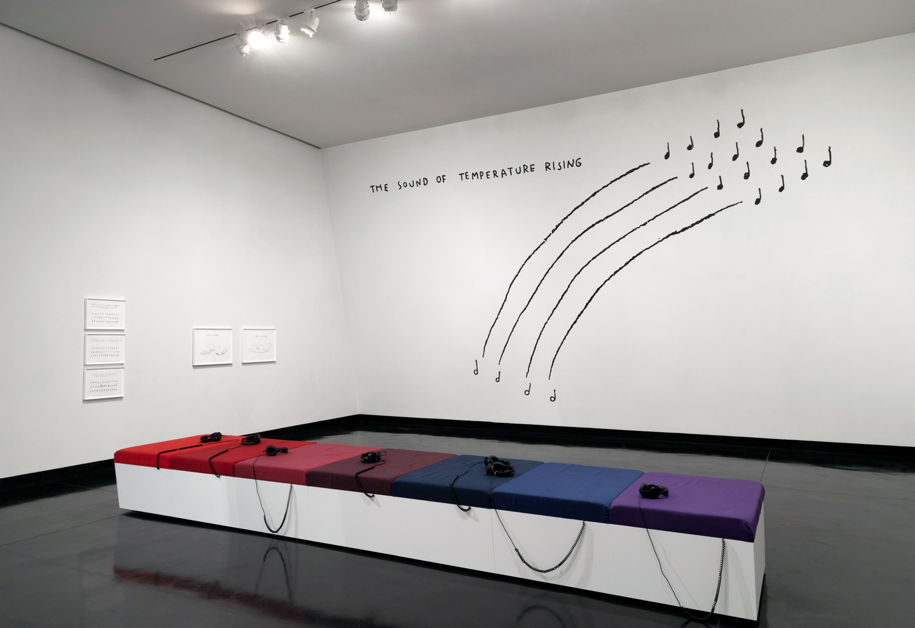 A gallery features one wall with five hanging artworks, and another wall with a large mural with black illustrations and text, and a bench with red, blue, and purple cushions.