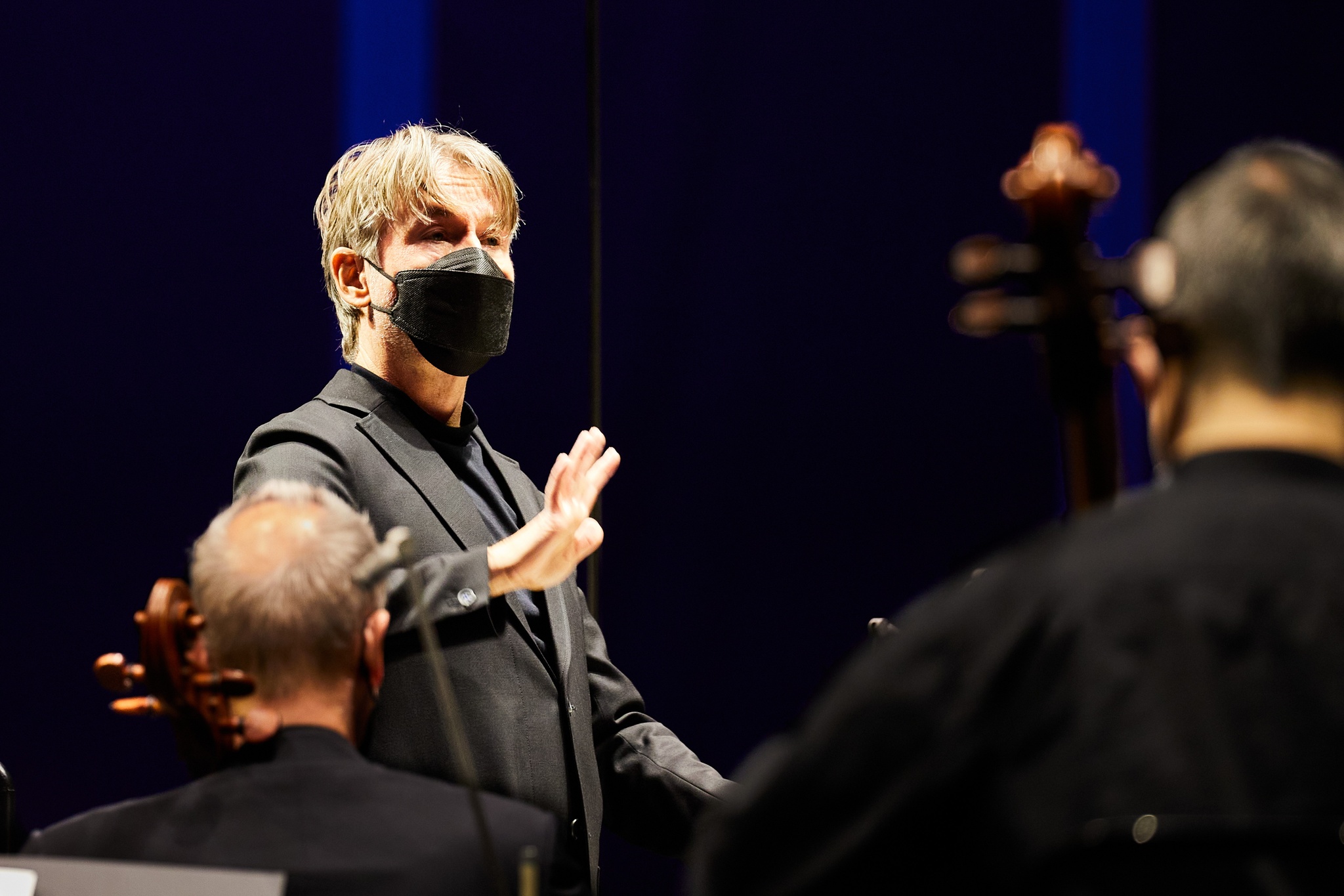 A conductor seen standing over the shoulders of orchestra members. He is a white man who wears a face mask and holds one hand up gesturing to the musicians.