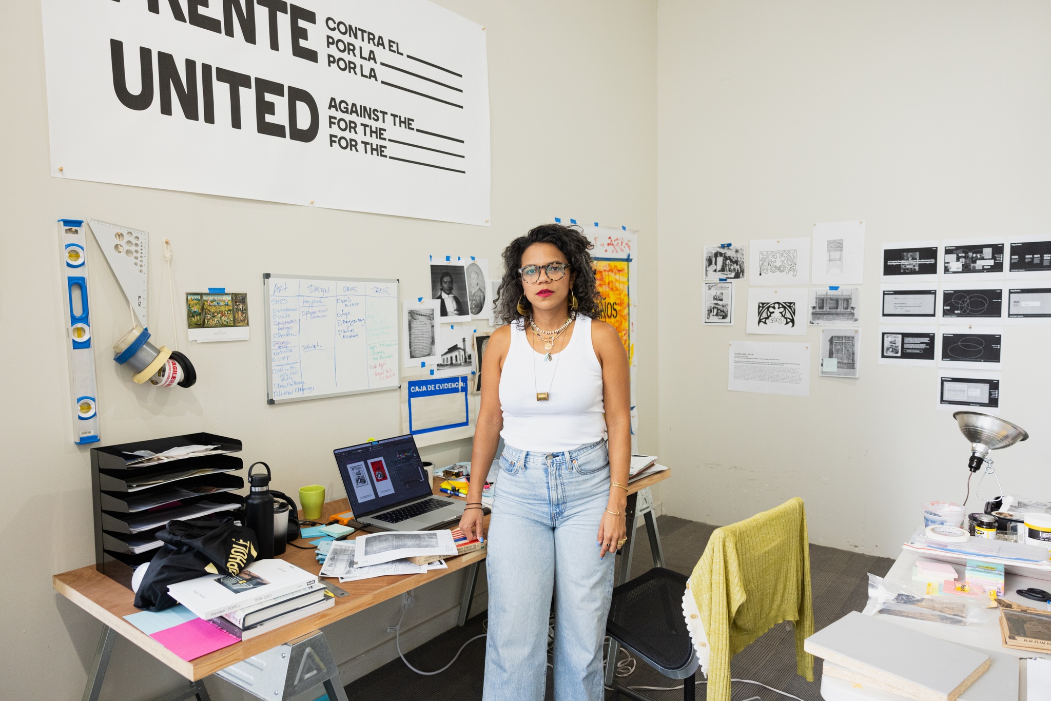 Dominican artist Lizania Cruz stands by a desk in her studio facing us. Lizania has wavy, shoulder length brown hair and wears glasses, a white tank top, and pale blue jeans. Her desk is covered with an open laptop computer and piles of papers. The walls behind her are covered in print outs and posters and sketches that are taped to the walls.
