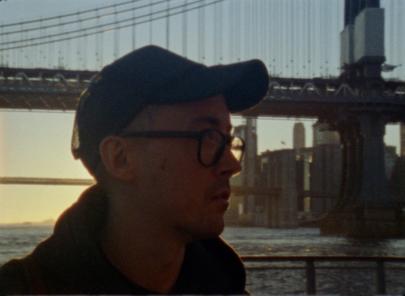 A portrait of Christopher Radcliff. He is seen in profile close up wearing a baseball cap and glasses. He is backlist by the setting sun behind which is setting behind the Manhattan and Brooklyn Bridges. 