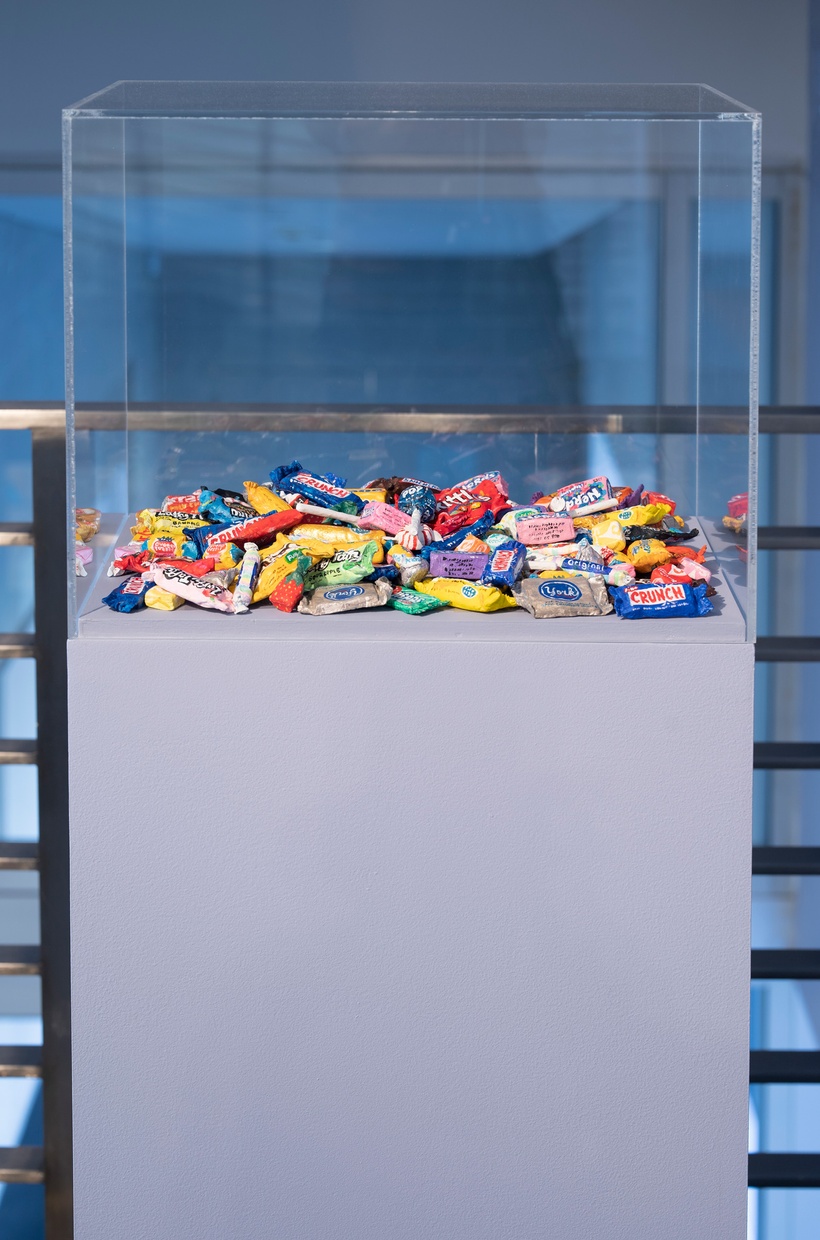 On a light purple pedestal within a square, glass case sit sculpted, colorful candies.