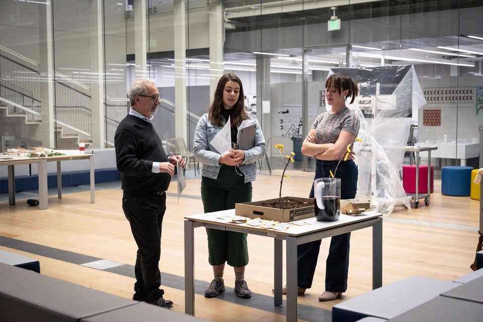 Three people stand around a table in a common space and discuss an artwork.