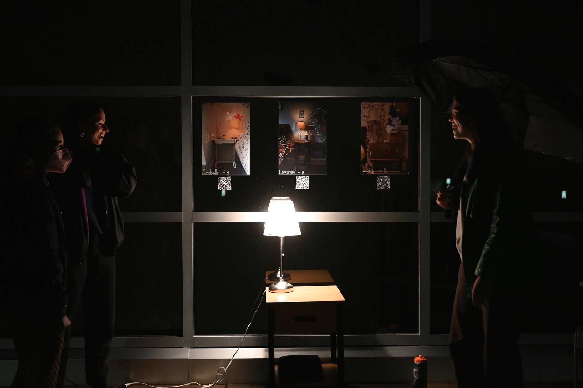In a dark space, three people look towards a bedside table with a lamp that sheds light on three artworks hanging above it.