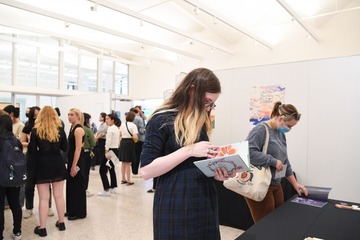 An exhibition space with multiple table booths. A student flips through a book by Kait Stansbury