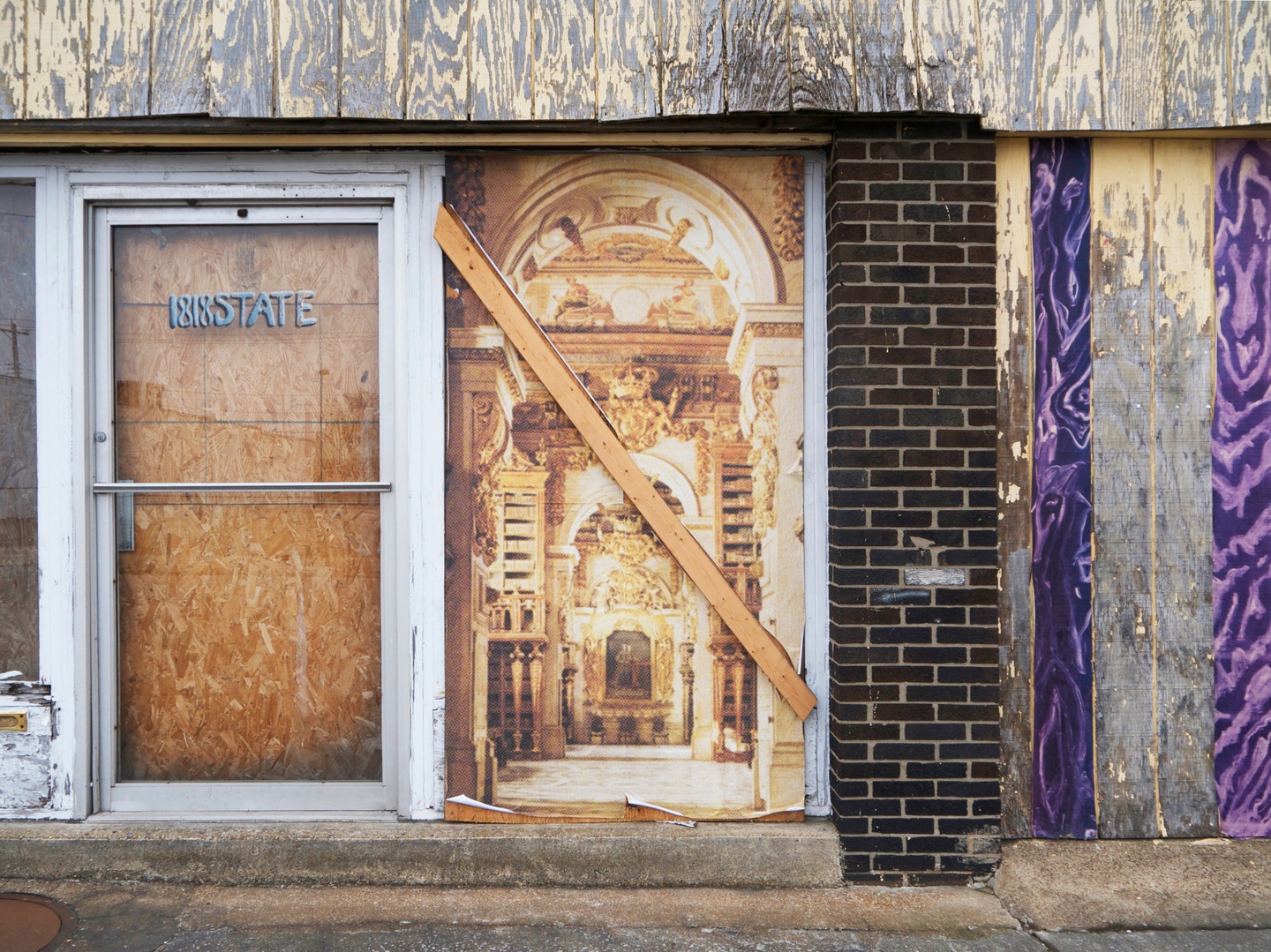 Picture shows an exterior wall of a building with a variety of different materials. From the left, a glass door shows plywood through the glass. The words "1818 State" are written on the door. To the right of the door is a printed board with an ornate space. To the right of that is a brick column, and finally there are set of painted boards.