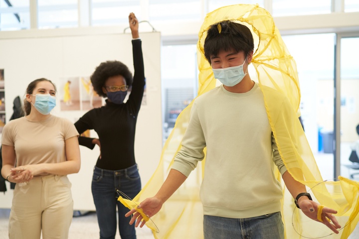 Three students in a gallery space, one with their hand in the air; the one on the right has a yellow garment draped over their head.