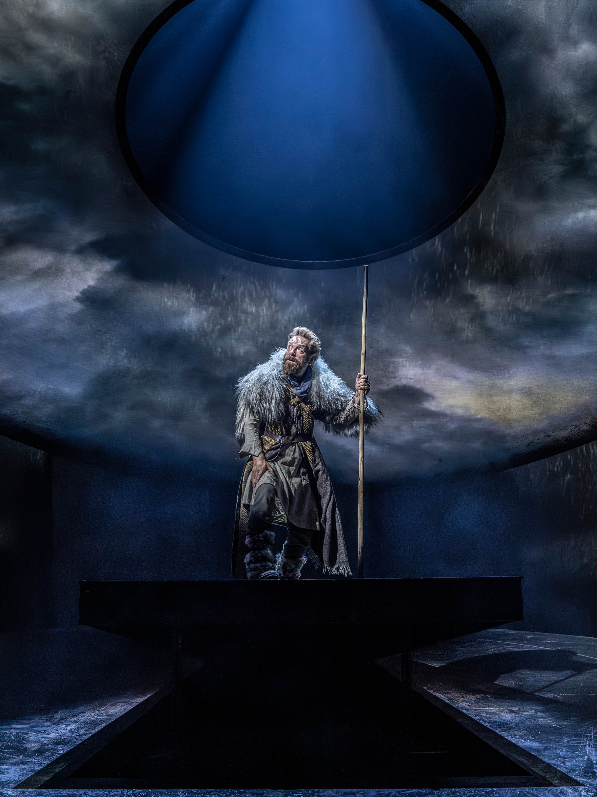 Actor Kenneth Branagh stands on a stage, atop a circular platform. We see him from an angle slightly below, looking up at him. He is dressed as King Lear with an animal pelt draped over his shoulders and holding a tall staff. Above him, a circular screen above the stage displays moody storm clouds. He turns his attention to the clouds with a look of wide-eyed trepidation on his face.  