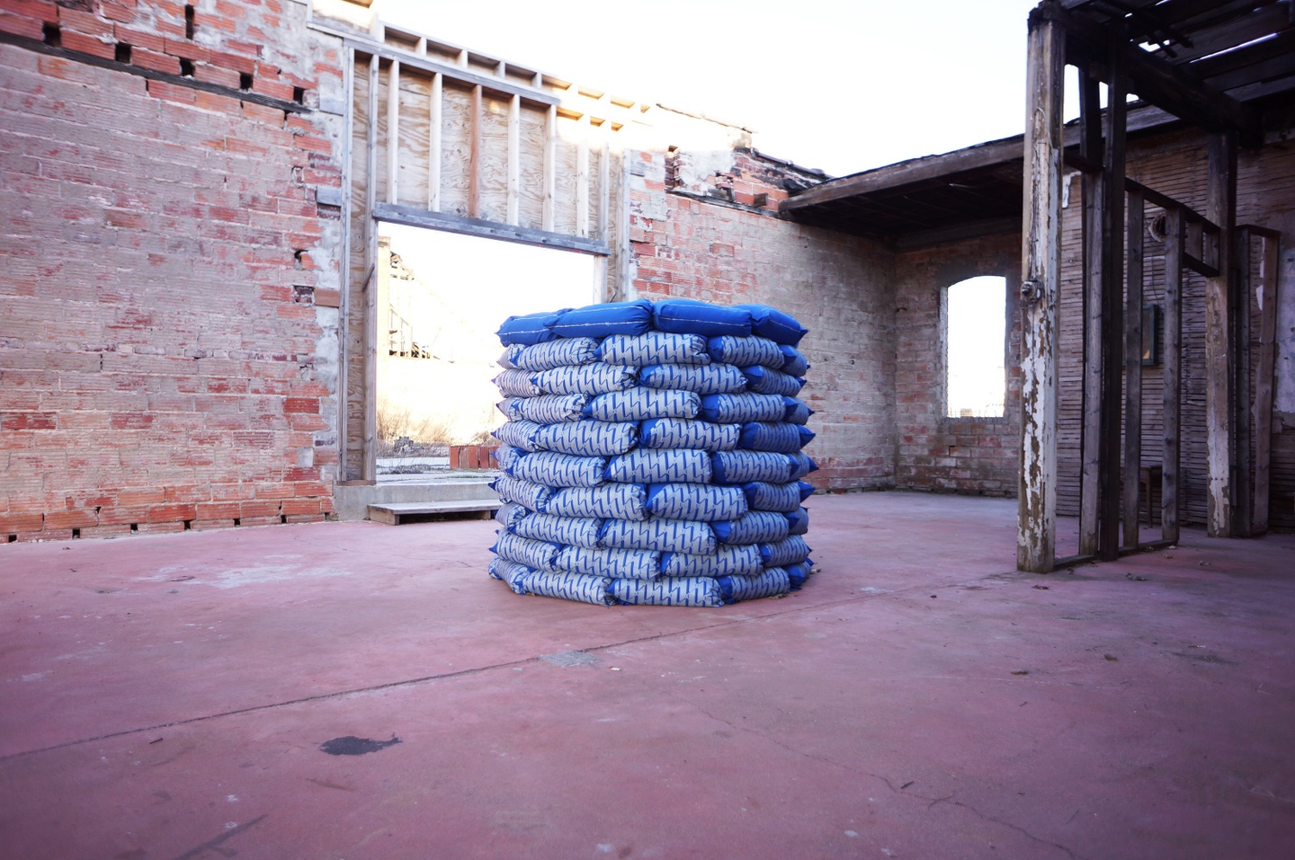 Photograph of a sculpture in space: the surroundings are an open-air brick and wood structure/building, with a faded red ground/floor; the sculpture is blue and white ,with a repeated geometric pattern printed on each layer.