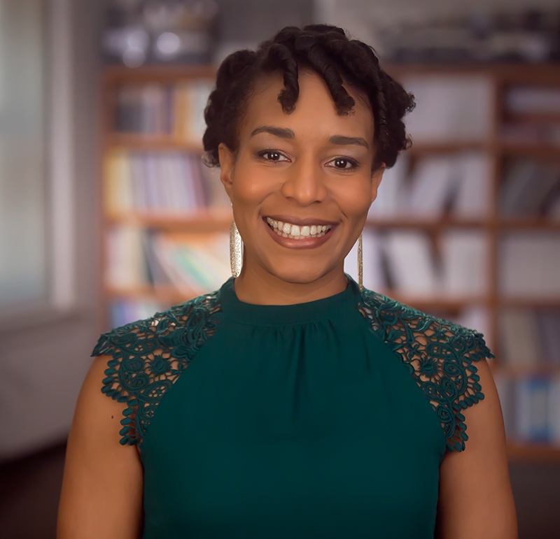 A photo of Barika X. Williams facing the camera and smiling. She wears a sleeveless green blouse with lace shoulders and a bookshelf lines the wall behind her in the out-of-focus background. 