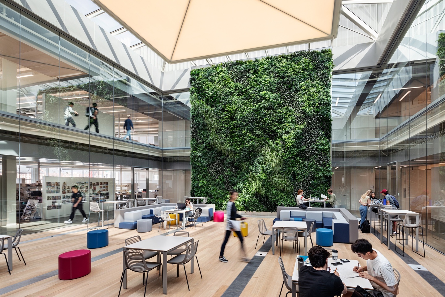 Interior 2-story tall courtyard with a skylight, a green wall, and colorful modular seating and tables, filled with students working and socializing.