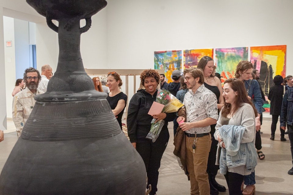 Three students stand in front of a black, 7 foot tall ceramic urn in a gallery space filled with visitors. Other paintings and sculptural artworks can be seen around the gallery.