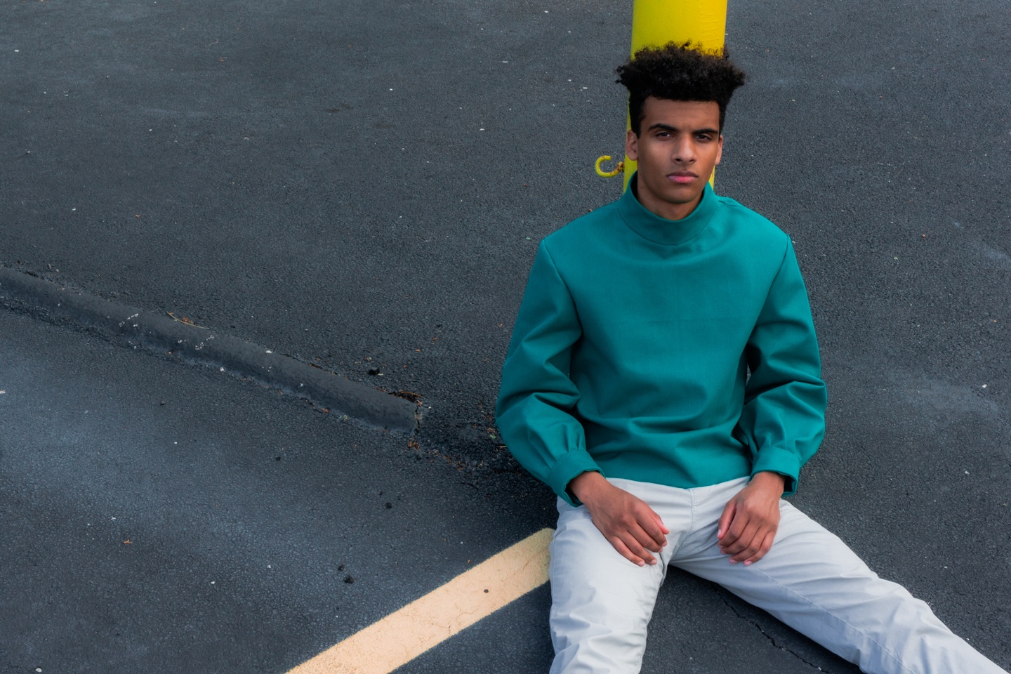 Model sits on an asphalt basketball court wearing white narrow legged trousers and a teal turtleneck with wide sleeves and tapered cuffs.