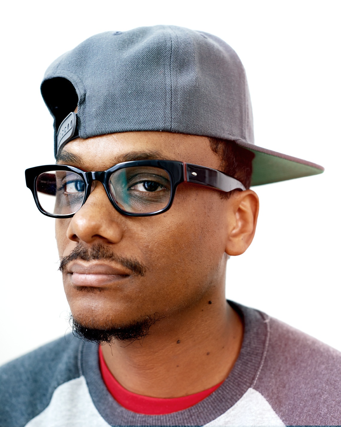 A portrait of DJ Donwill. Donwill is a Black man who wears a backward baseball cap with a straight bill, rectangular glasses, and a t-shirt with a red undershirt visible just under the neck.