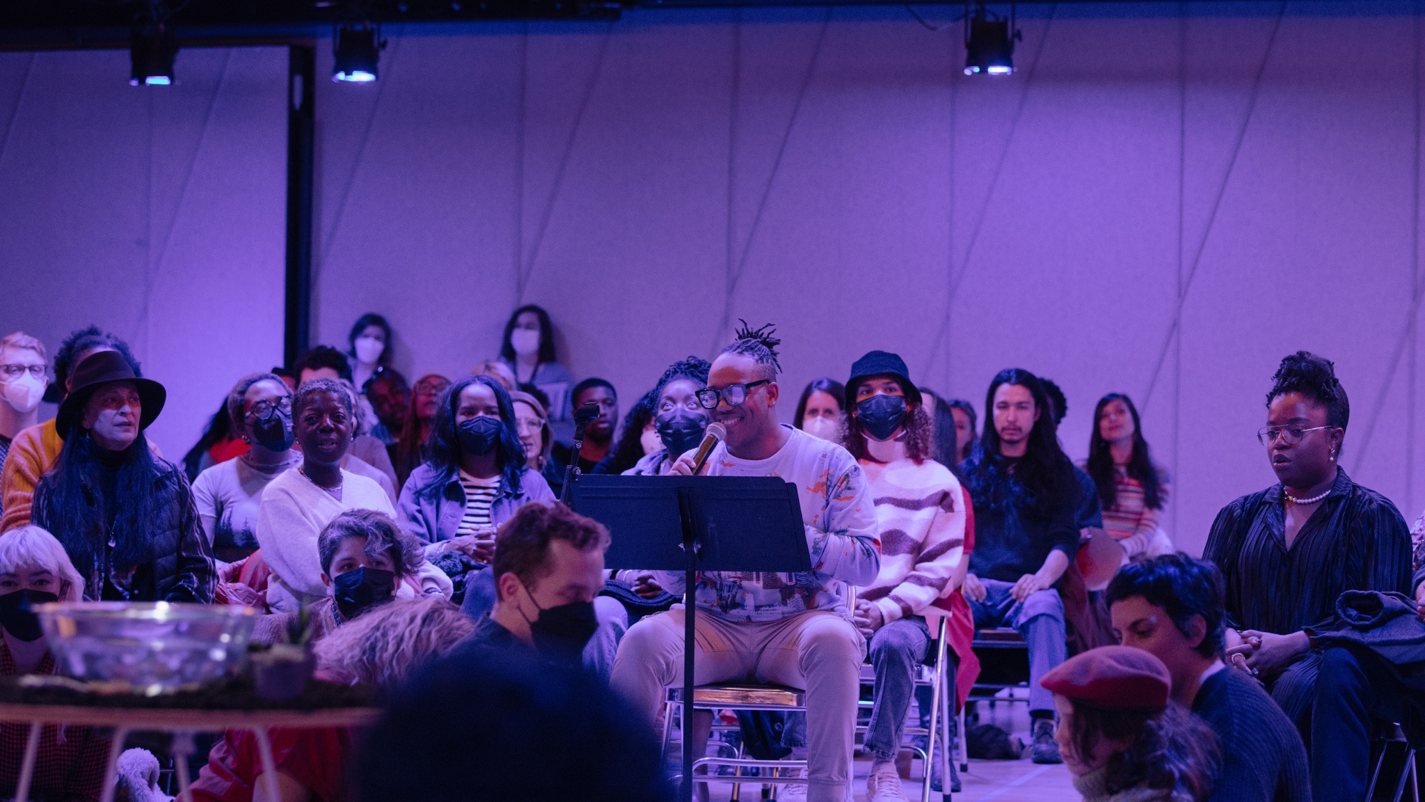 A performance space suffused in purple light. Choir members sit in chairs arranged in multiple rows in a circle with audience members mixed in. At the center, Troy Anthony, a Black man and artistic director of The Fire Ensemble, speaks into a microphone.  