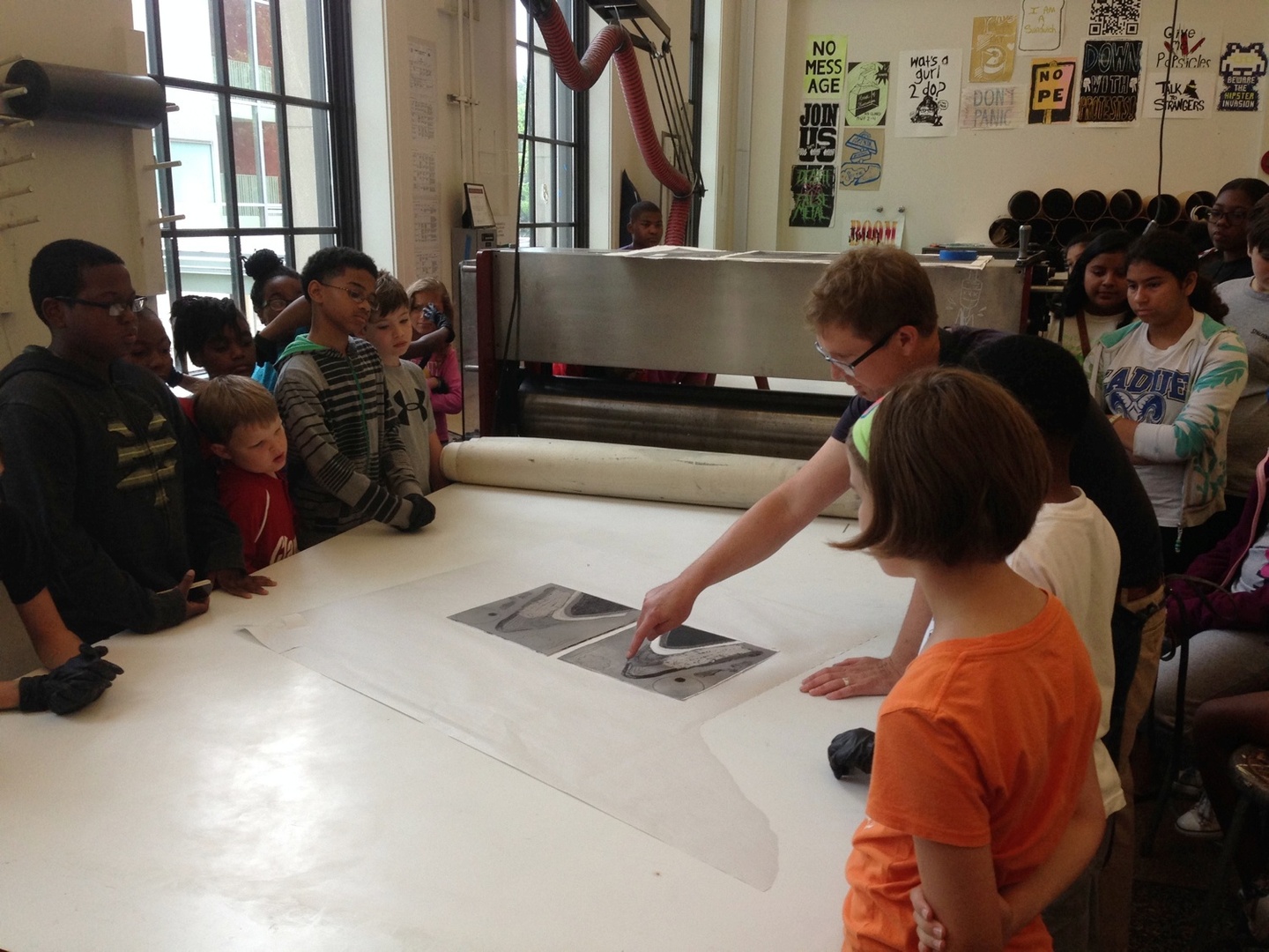 A group of children look at a person pointing at a printing press.