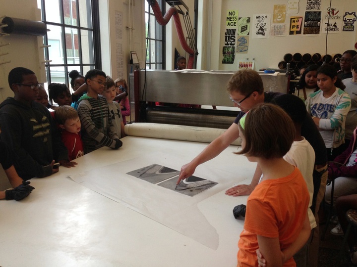 A group of children look at a person pointing at a printing press. 