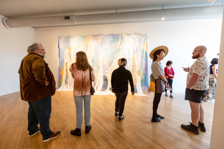 Gallery view with visitors in front of Anna Schenker's large multicolored print overlaid with cut out pieces of tree rubbing print hanging on a wall