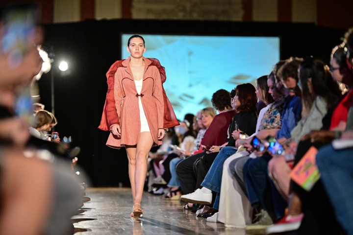 A model walking down the runway in a pink salmon-colored jacket with swirl mesh details