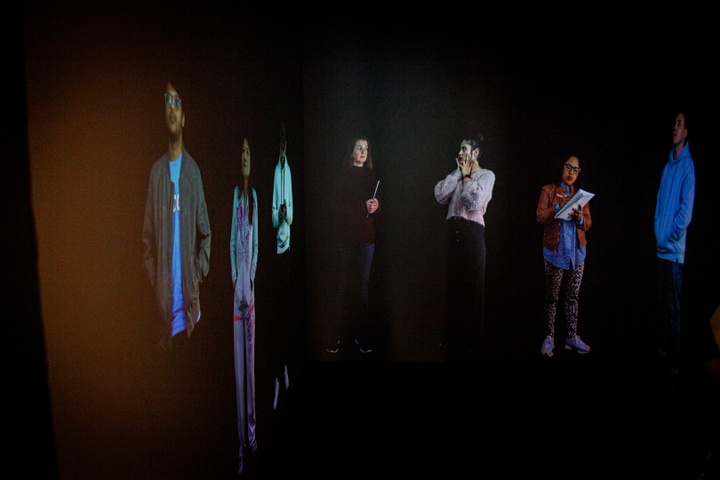 Black box room with life-size projections of people of various ethnicities standing and looking around them.