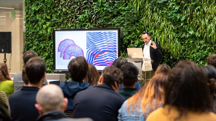 An individual standing in front of a lush green wall of plants delivers a presentation to a seated crowd. A digital monitor featuring architectural work is to his side.