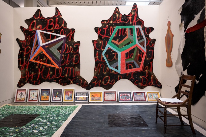 Studio space with the back wall completely covered by two unevenly shaped canvases with black backgrounds. Red cursive letters of the alphabet form the background and depictions of an octahedron and a dodecahedron in the foreground. A line of smaller paintings is stacked along the bottom of the wall.