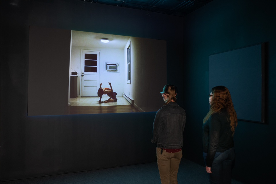 Two people in facemasks stand together in a black box room to view a projection. The video still is of a person in a contorted pose in a small kitchen space.