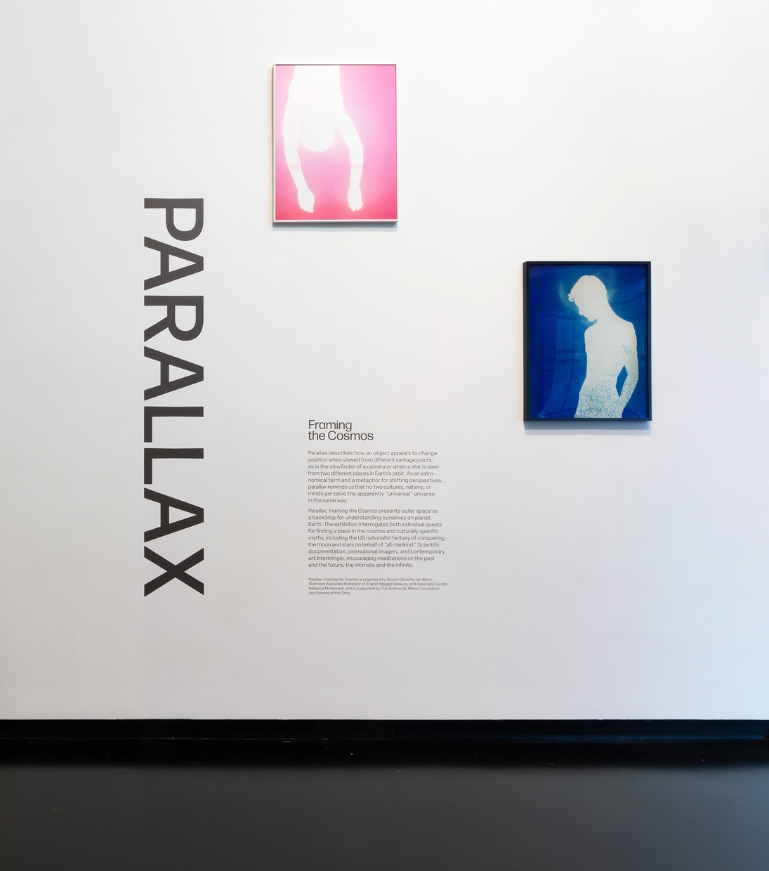 A white wall features two hanging framed artworks and large, black, vertical text that reads “PARALLAX,” to the left of two paragraphs of smaller black text.