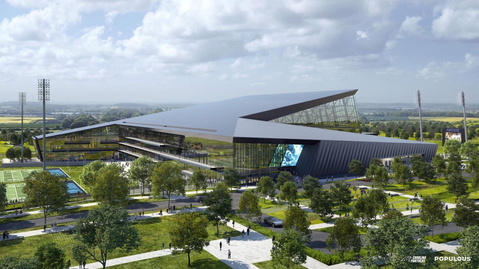 Exterior facade of the Carolina Panthers Training Facility and Headquarters. The irregularly shaped building has a gray roofline that branches off to two triangular sections. Lots of windows are featured on the facade of the multi-story building, which is surrounded by trees and walkways, as well as football practice fields to the left.