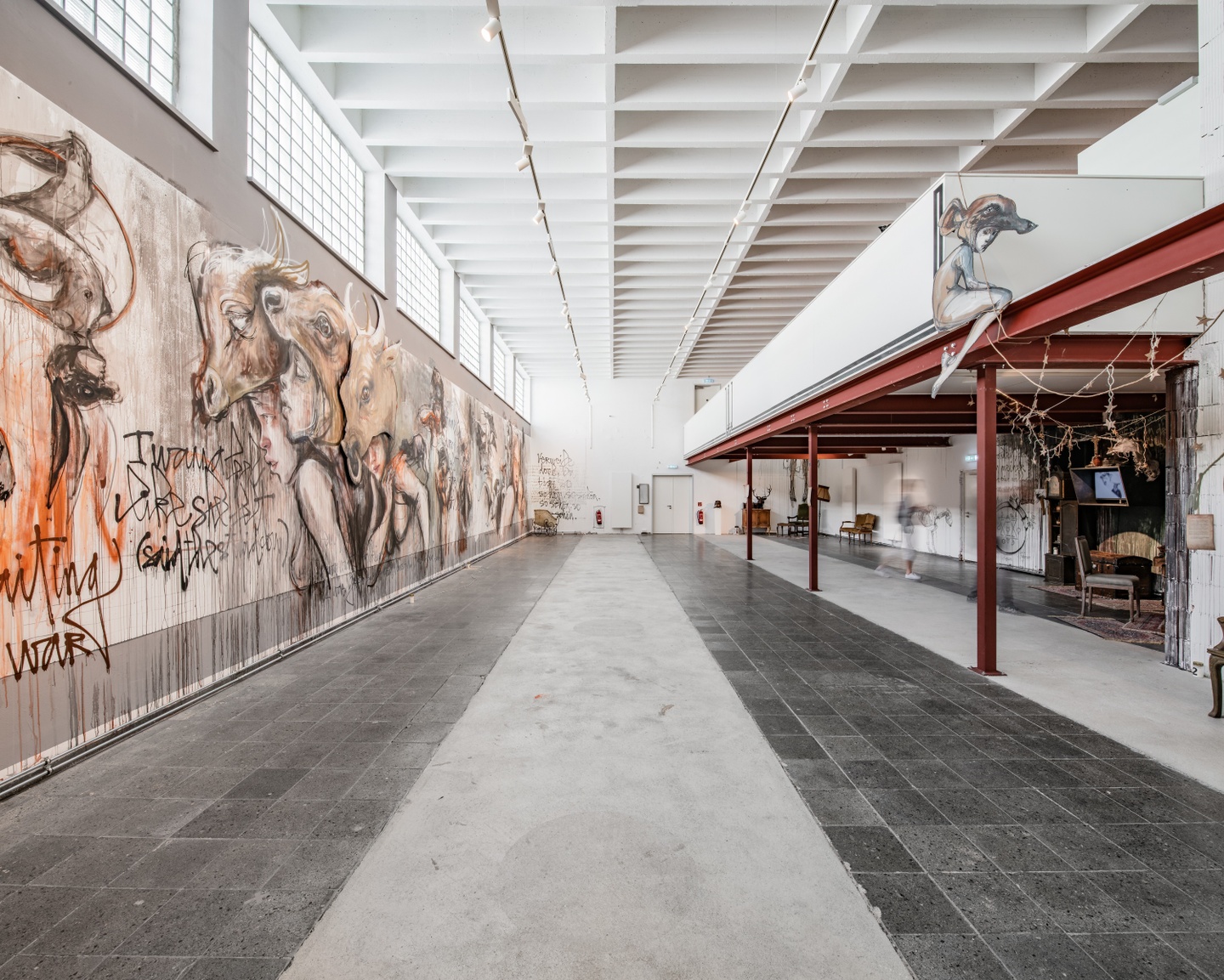 Interior of a museum space with industrial-windows and an iron framework. One wall displays a large-scale painting that is a hybrid of graffiti and cave painting.