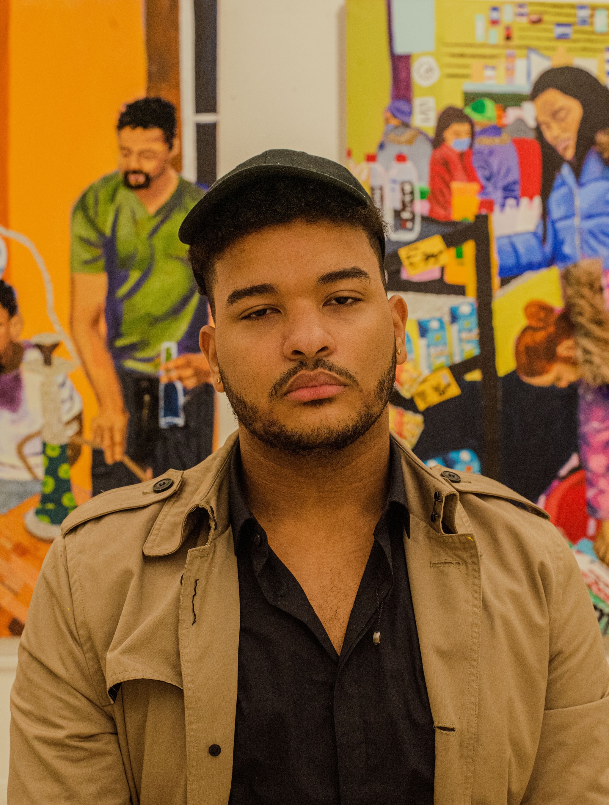 A headshot of Bryan Fernandez, a Dominican artist. He wears a dark cap and a tan jacket over a black shirt. He has a mustache and trimmed beard and poses in front of a painting. Photo by Kiara Terrero