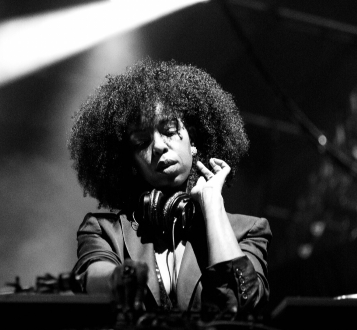 A black-and-white portait of DJ Reborn at a turntable. She is a Black woman wearing a jacket with lapels and holds a a pair of headphones around her neck with one hand.