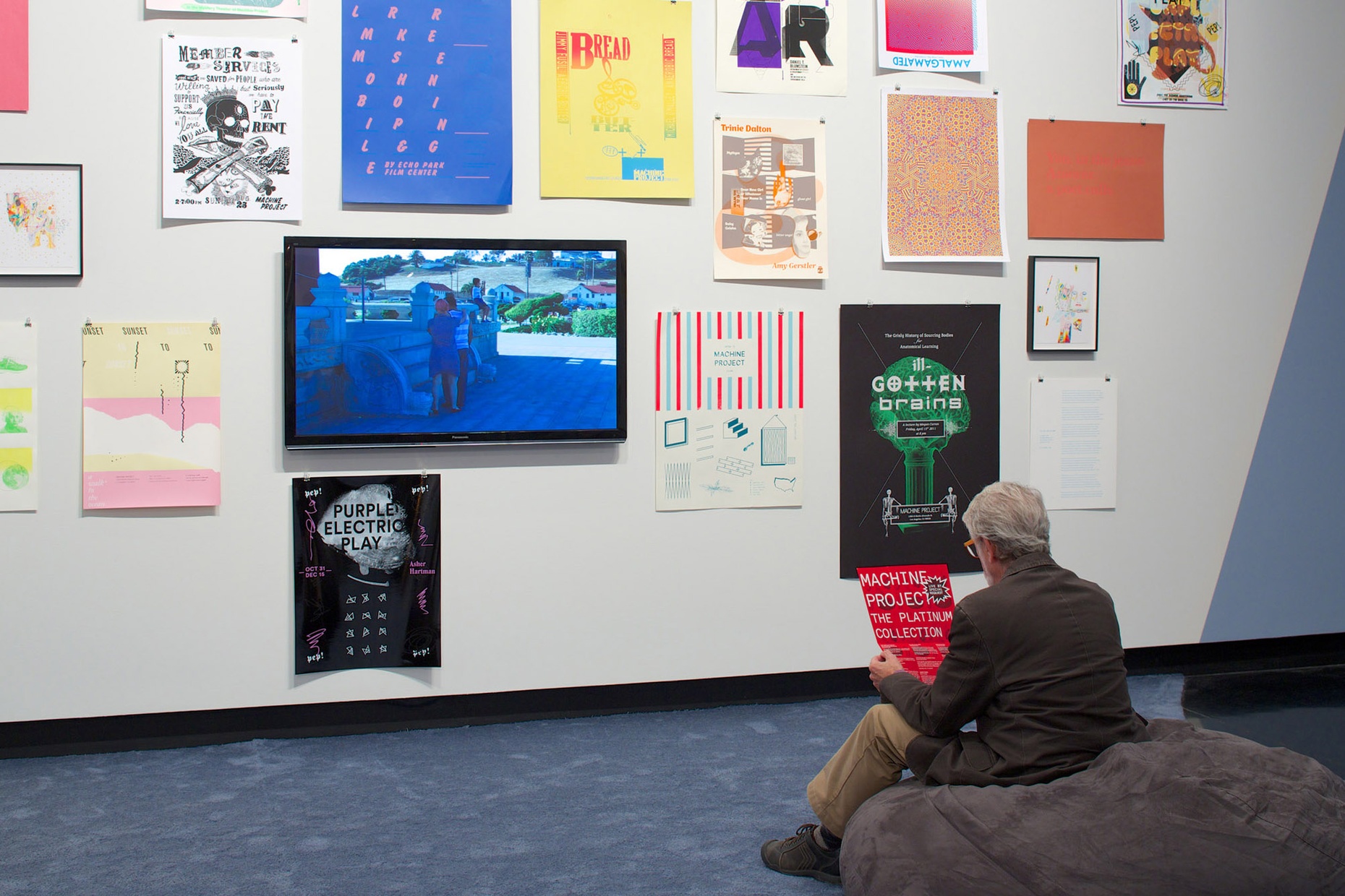 A man sits on a large bean bag chair holding a poster and facing a wall of posters.