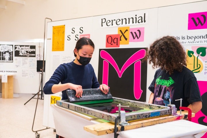 Two people work a screenprinting stand in a gallery space displaying typographic posters.