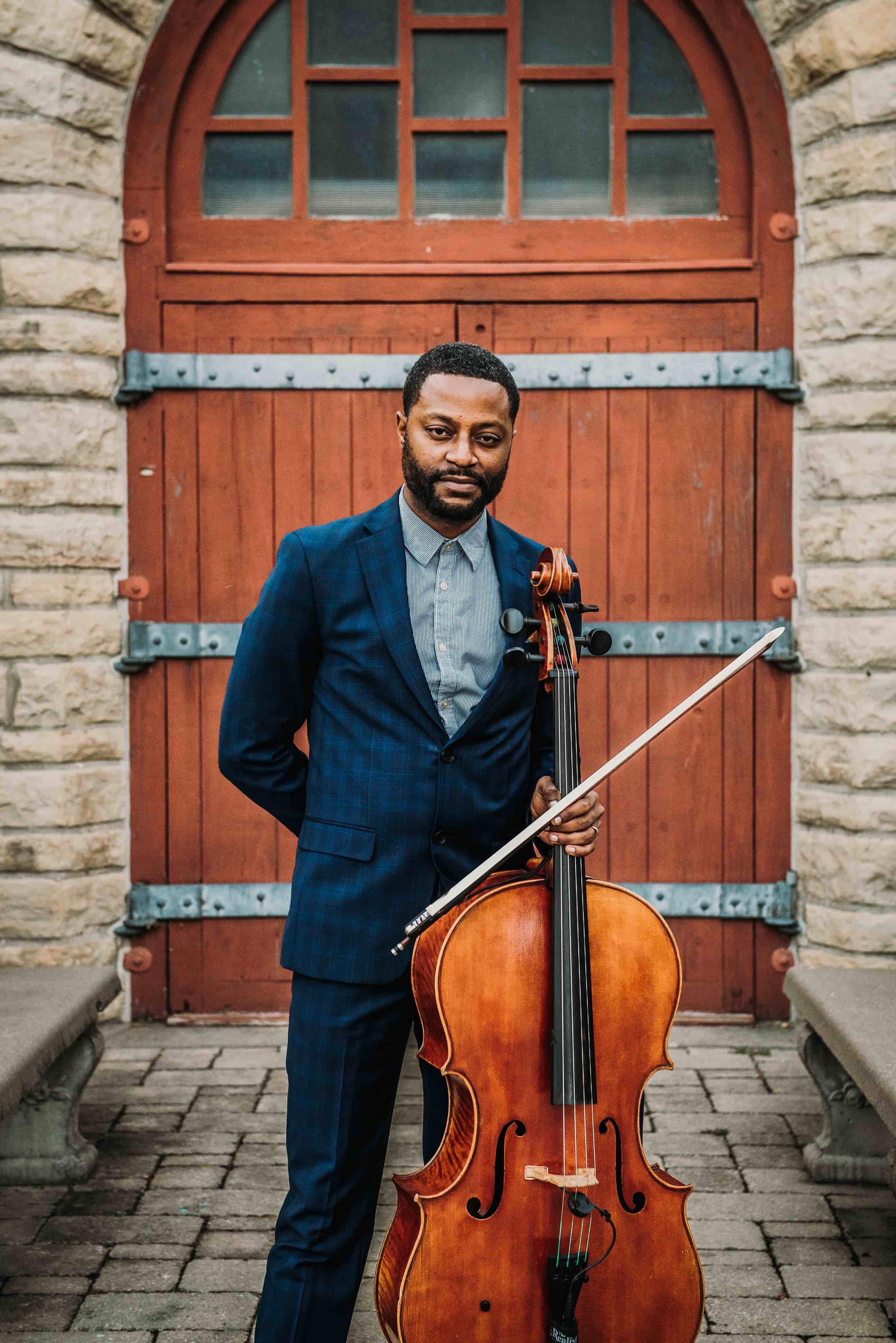 A Black man in a blue suit standing with a cello and bow in front of wooden doors topped by windows in an archway. 