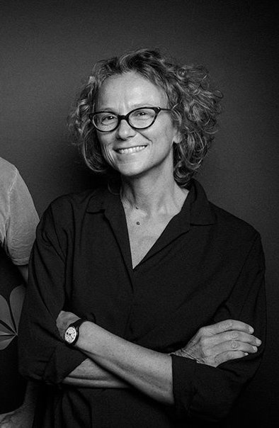 A black-and-white photo of Julia Solomonoff smiling with her arms crossed over her torso. Solomonoff wears glasses with slightly oblong frames around the lenses. 