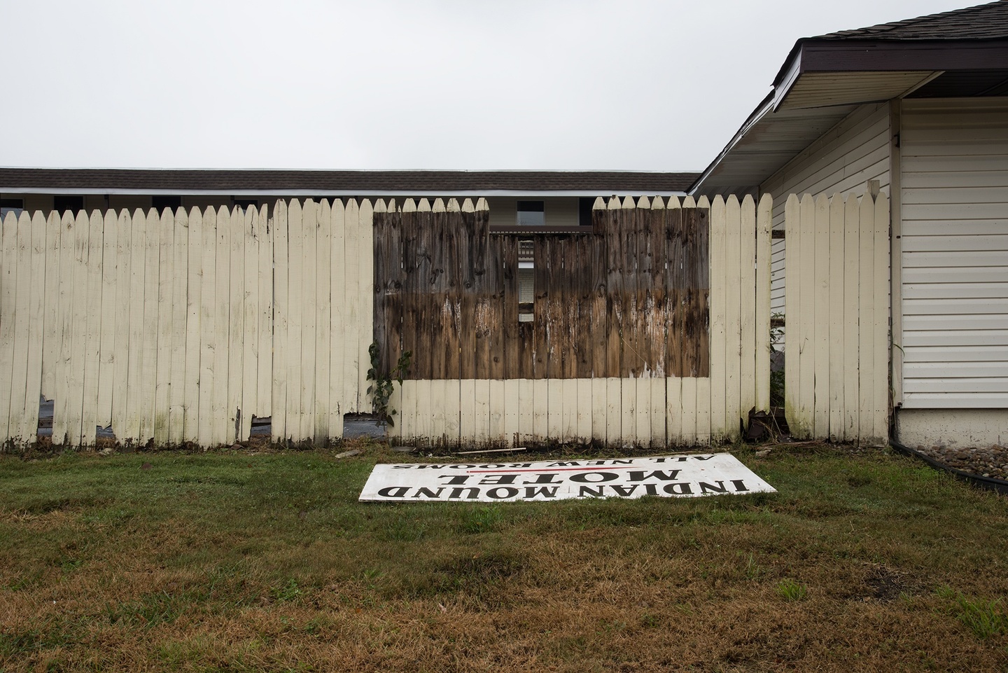 A light-hued fence along the side of a house: a rectangular portion is a darker brown, in front of which on the ground is a hand-lettered sign that reads, "INDIAN MOUND MOTEL" — "ALL NEW ROOMS." The sky is white and the grass on which the sign rests is green-brown.