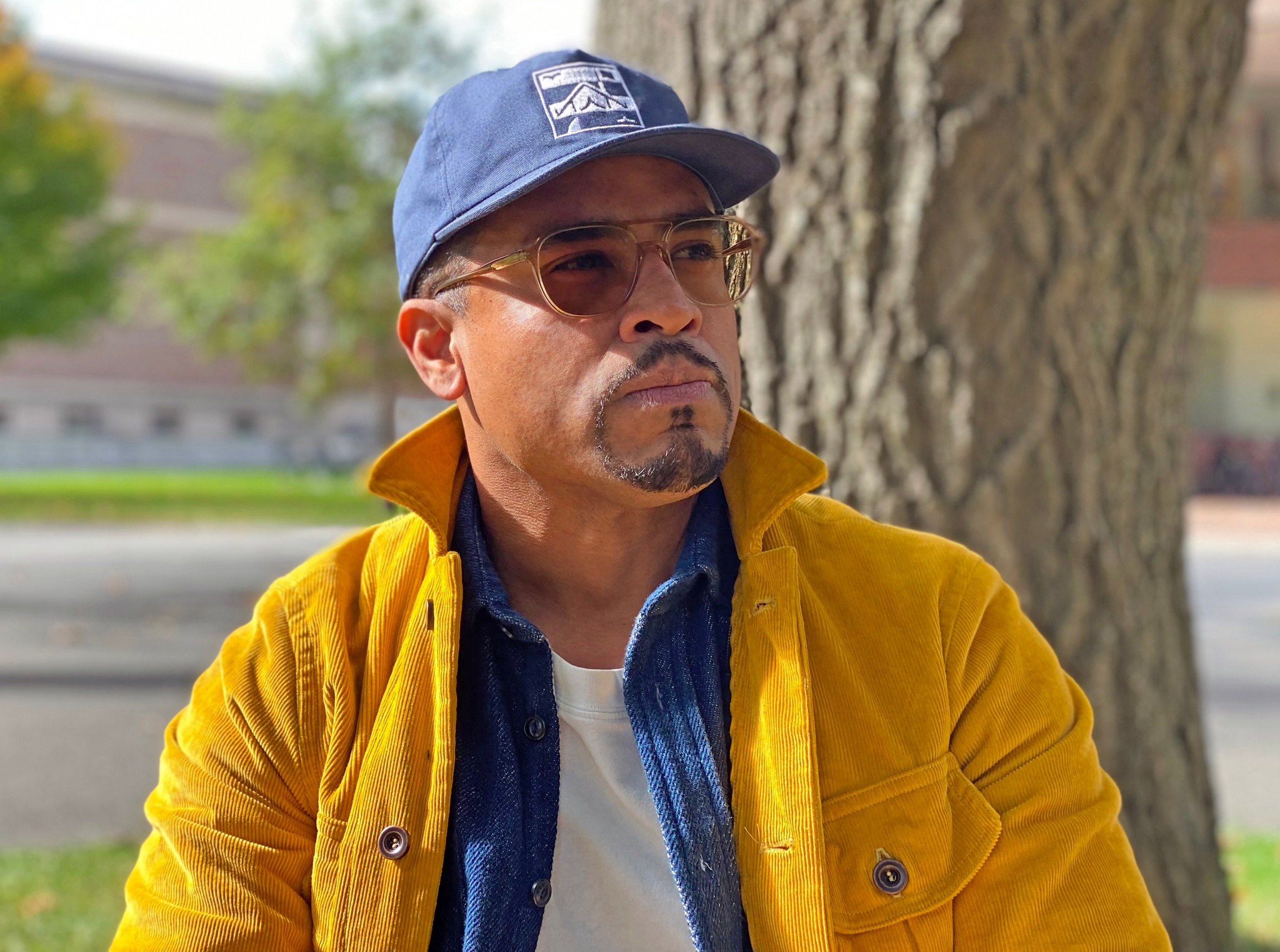 A portrait of Shaun Leonardo outside posing against a tree. He wears a blue basseball cap, sunglasses, and a yellow jacket. He looks off to the side.