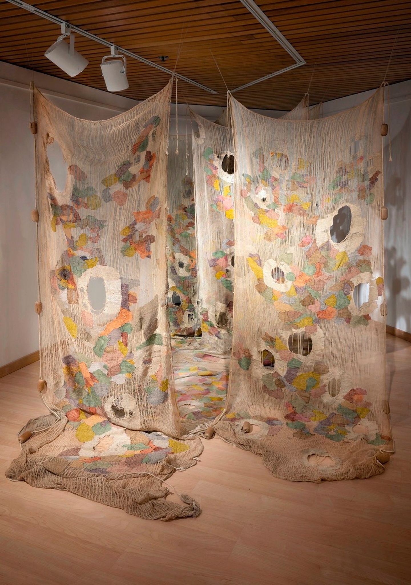 4 large pieces of fabric hanging from the ceiling and down to drape on the floor in a gallery space, Each piece is beige with large random holes and patches of colors sewn on