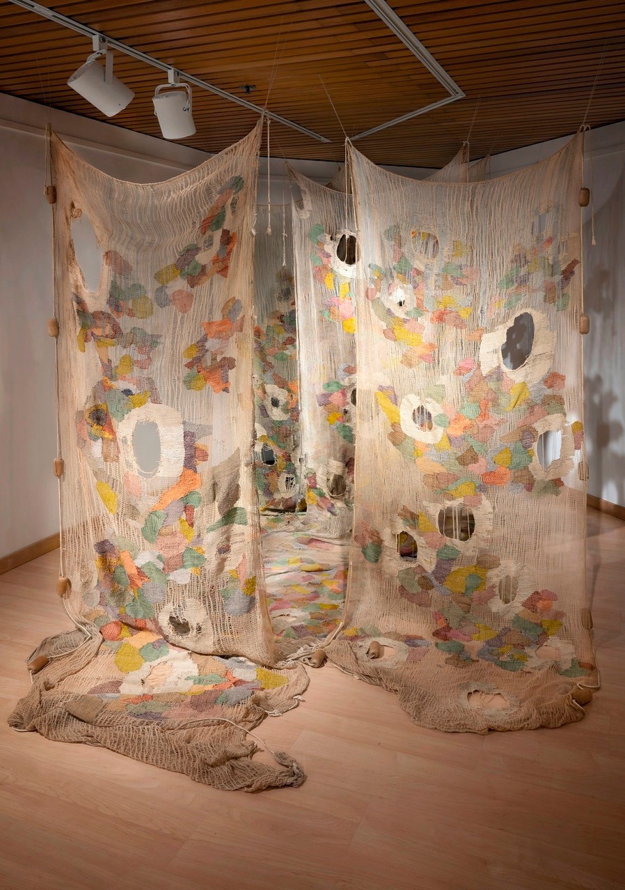 4 large pieces of fabric hanging from the ceiling and down to drape on the floor in a gallery space, Each piece is beige with large random holes and patches of colors sewn on