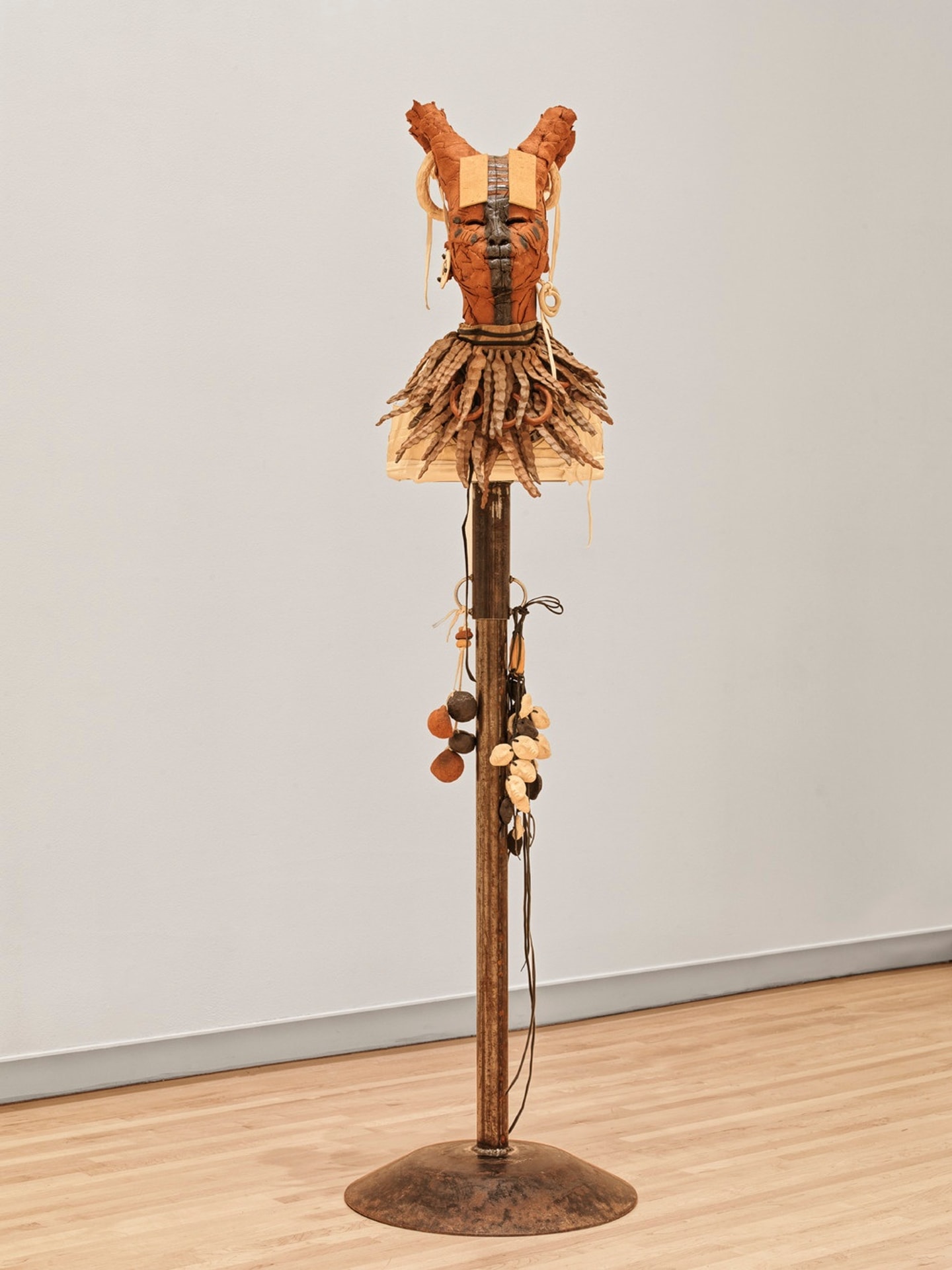 Sculpture of a stylized human figure with a terracotta-colored head, horn-like protrusions, and a layered, textured collar, mounted on a tall wooden pole with dangling cords and objects, displayed on a gallery floor.