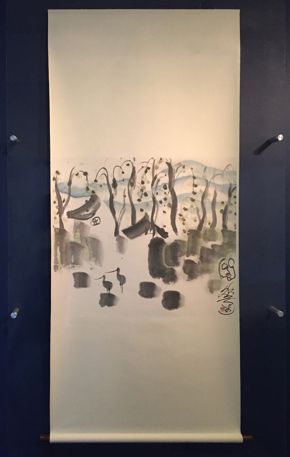 A long tan paper scroll with an ink painting of two herons in a simple village-scape. 