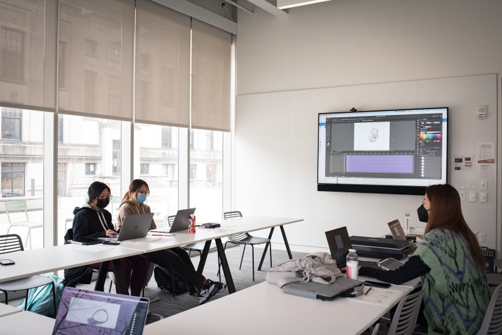 Several people with laptops sit at a U-shaped conference table around a video screen displaying an animation timeline.