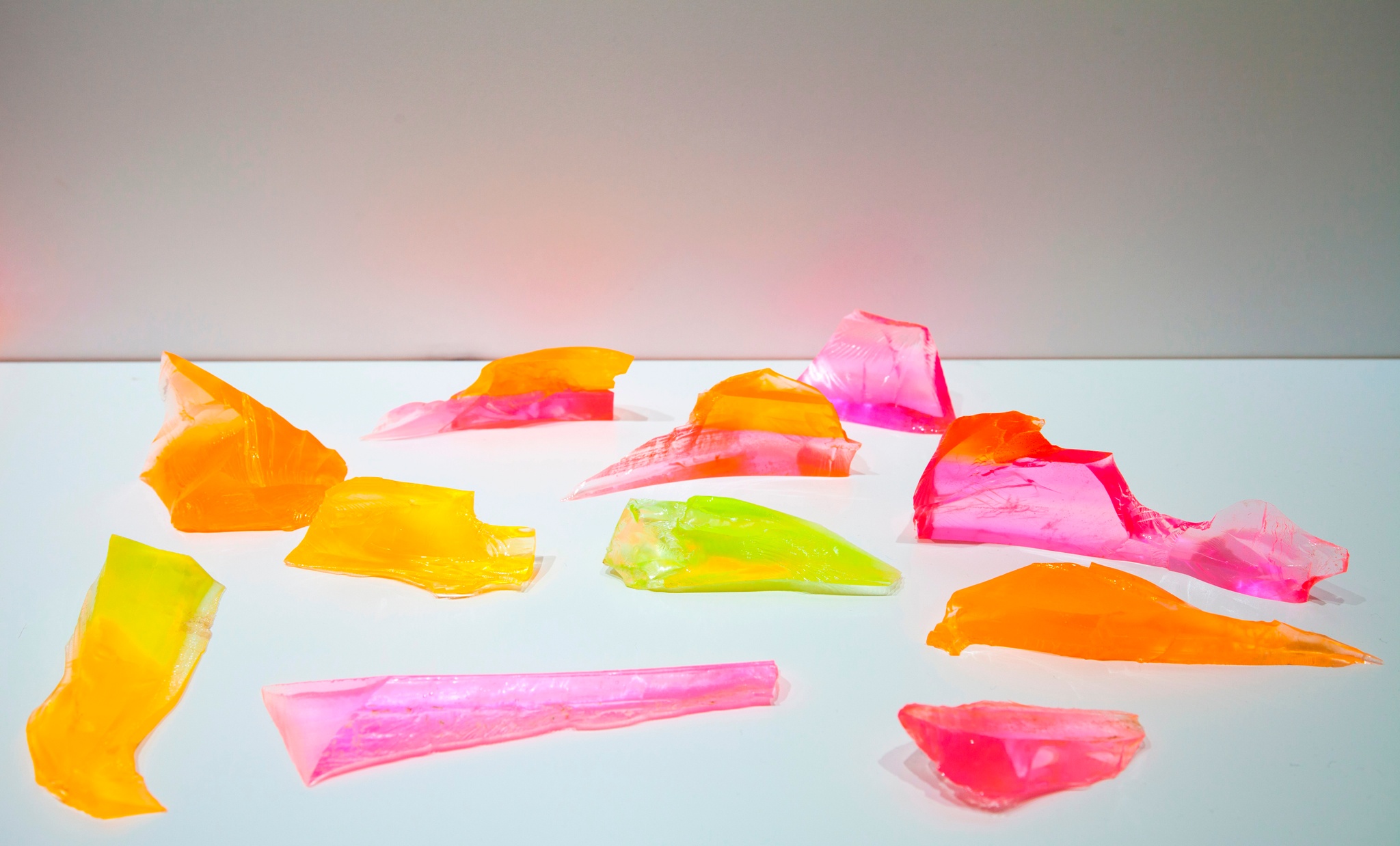 Transparent fragments, as if a glass has been shattered, are scattered on a white surface. The shard-like fragments are fluorescent pinks, oranges, greens, yellows, and purples.
