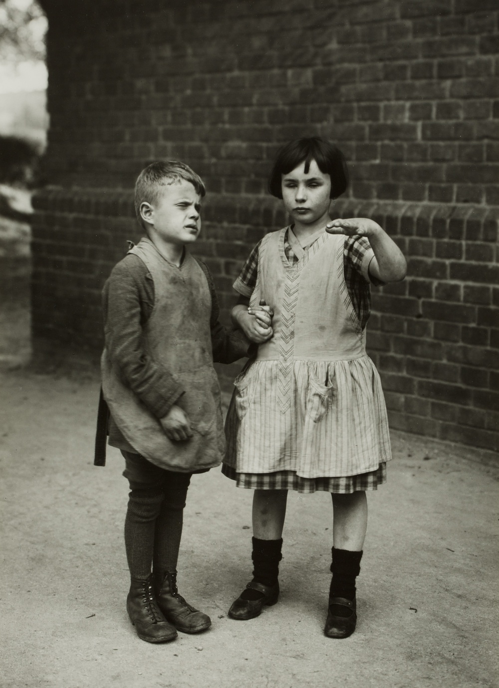 A black and white photograph of a young boy and girl standing in the street holding hands and wearing dirty clothing. The girl’s hand is suspended in front of her body at shoulder height.