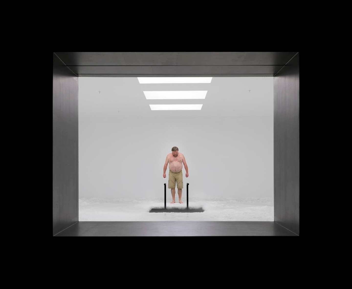 A black frame around a view into threshehold of a white room with three rectangular, overhead lights. A shirtless individual is looking down at a rectangular opening in the floor, with what might be a black ladder leading into it.