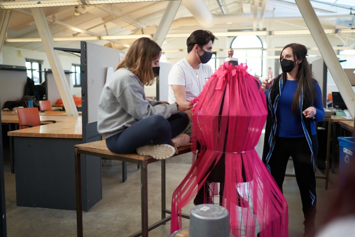 Two students sit on a work table behind a magenta fabric construction on a mannequin. An instructor comments on the project.