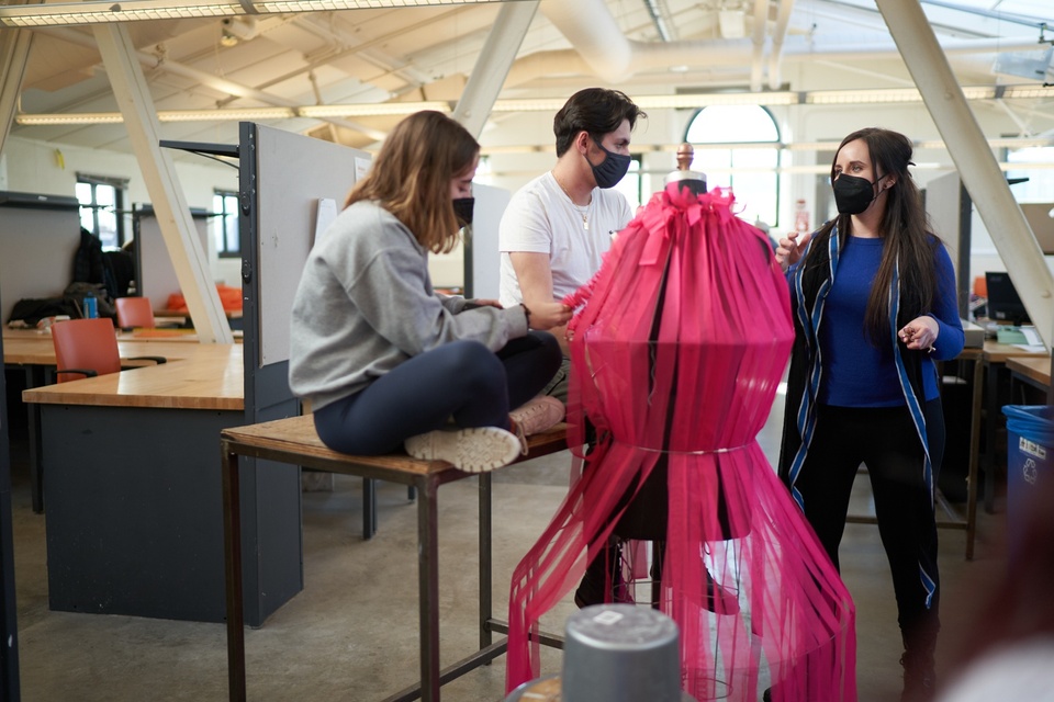 Two students sit on a work table behind a magenta fabric construction on a mannequin. An instructor comments on the project.