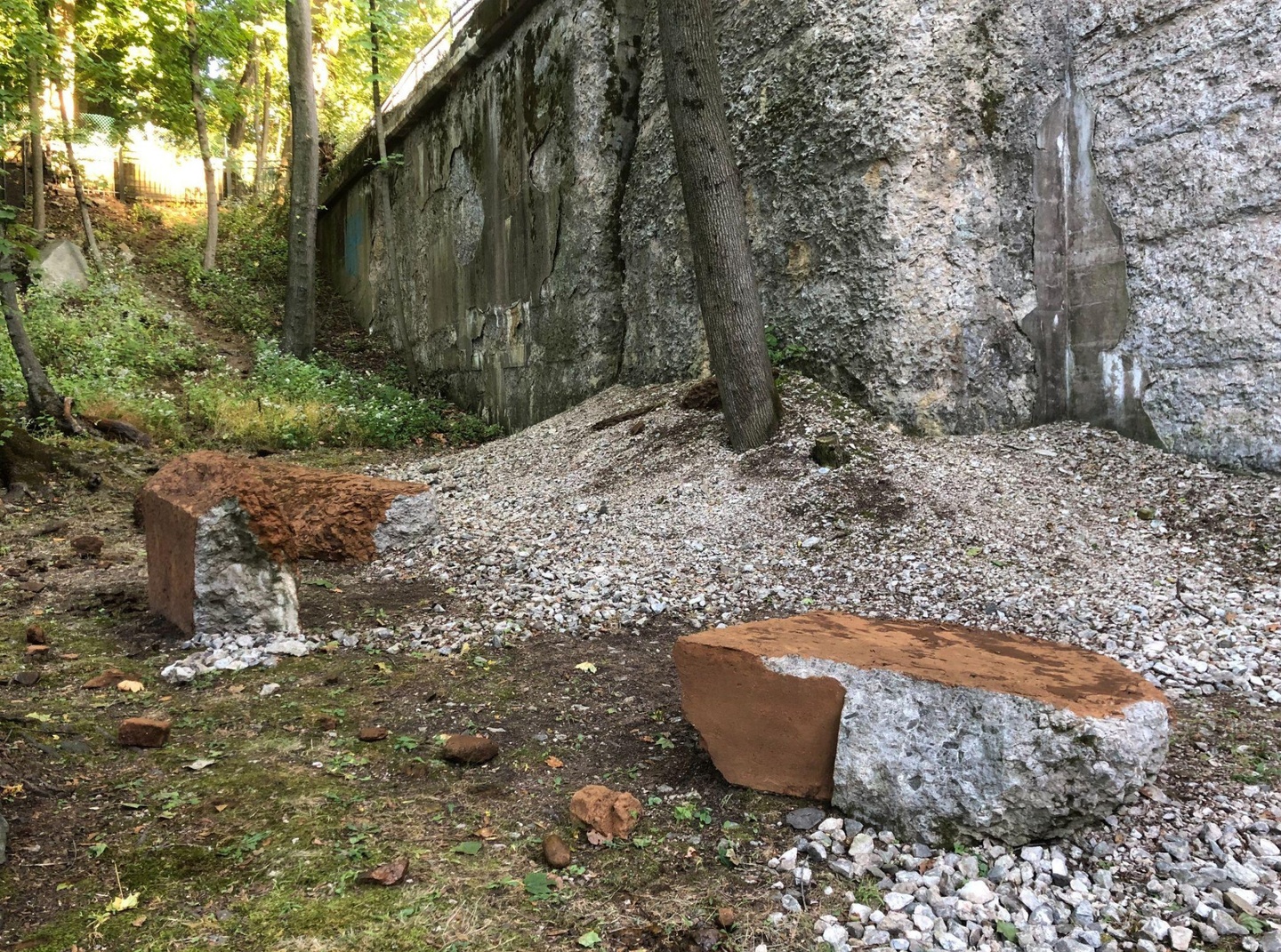Sculptural installations of concrete and architectural fragments, set outdoors between a wooded area and an old wall.