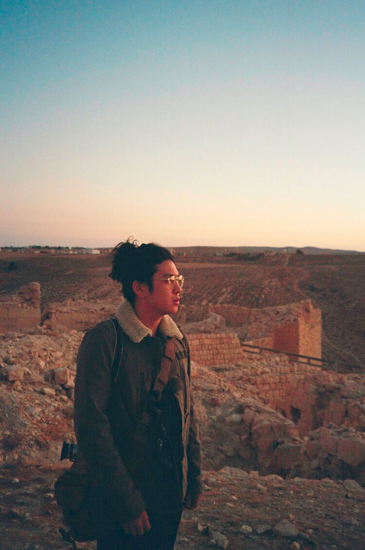 A young Chinese American man stands amidst a desert-like background with structures that look like ruins partially in the earth. He wears glasses, a warm jacket with a camera strap across his chest, and has long hair gathered up in a loose bun.