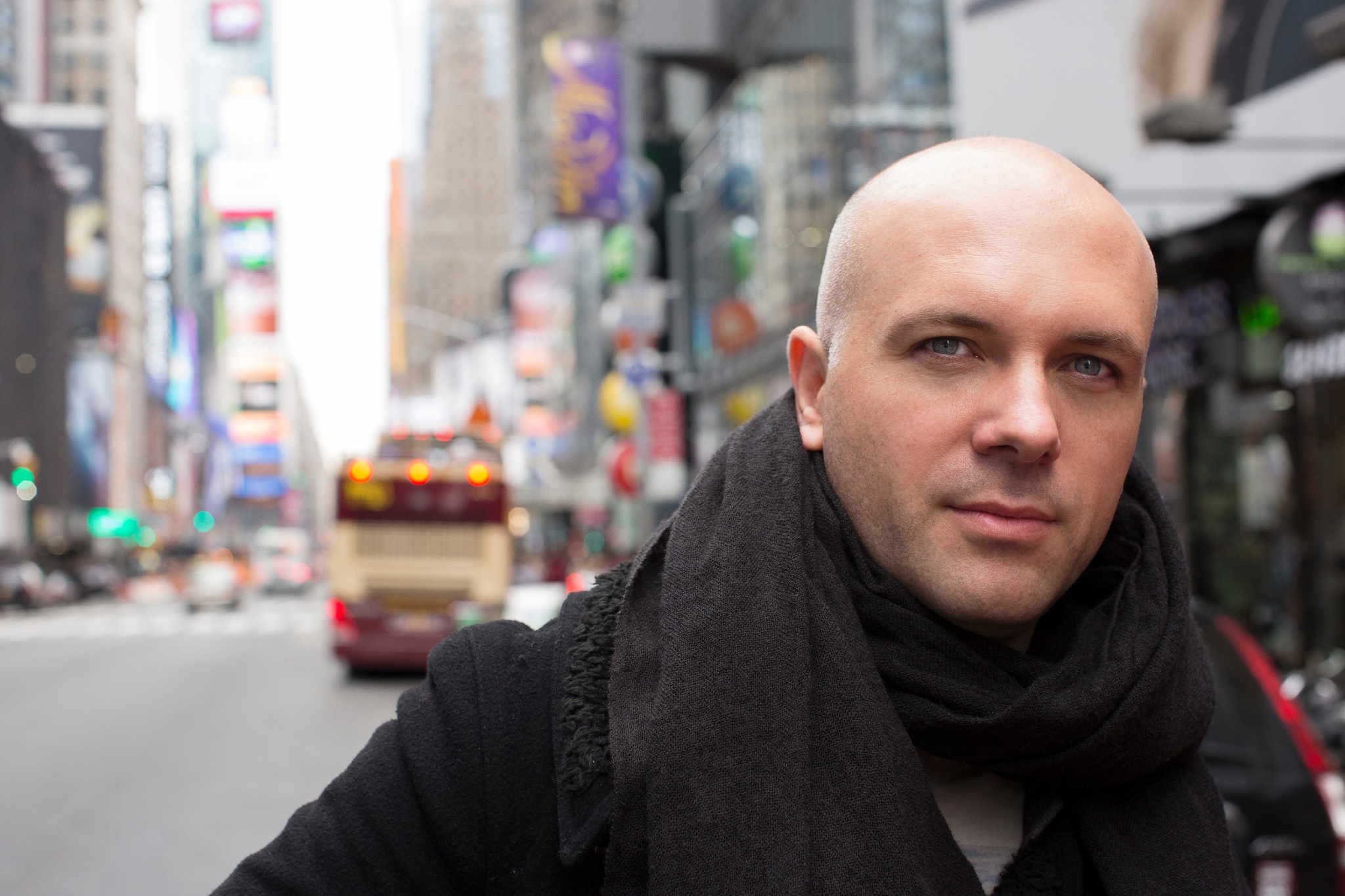 A white man with a bald head seen from the shoulders up standing in city with a street with buses and cars and lined by tall buildings receding behind him. 